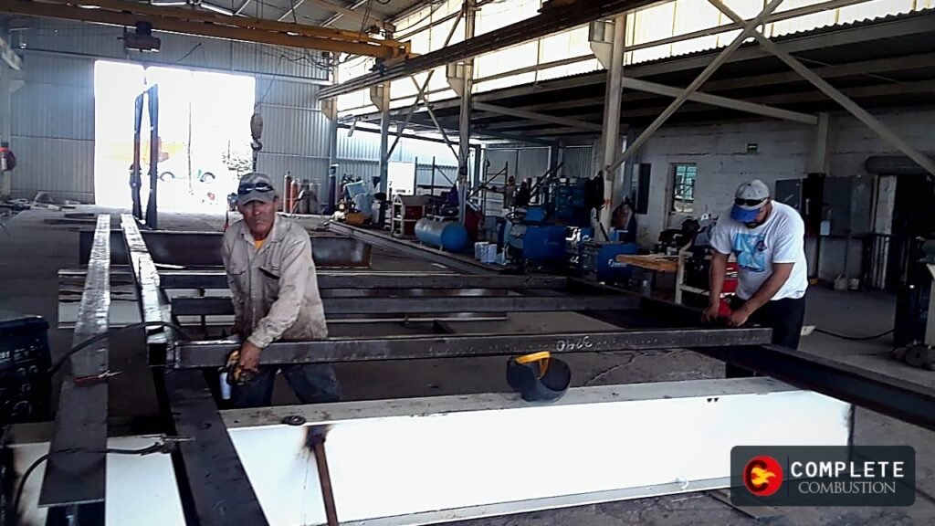 Workers assembling a large steel frame in a fabrication shop, with Complete Combustion logo in the bottom right.